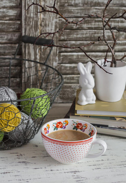 舒适的家庭座椅，有一个带纱线的篮子，堆叠的书，有干树枝的花瓶，一只陶瓷兔子和一杯带牛奶的茶。