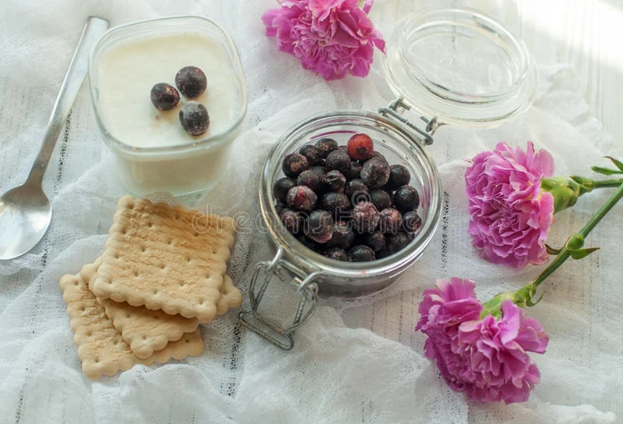 一罐新鲜浆果一杯自制酸奶饼干和明亮的紫色花朵在精致的白色织物表面