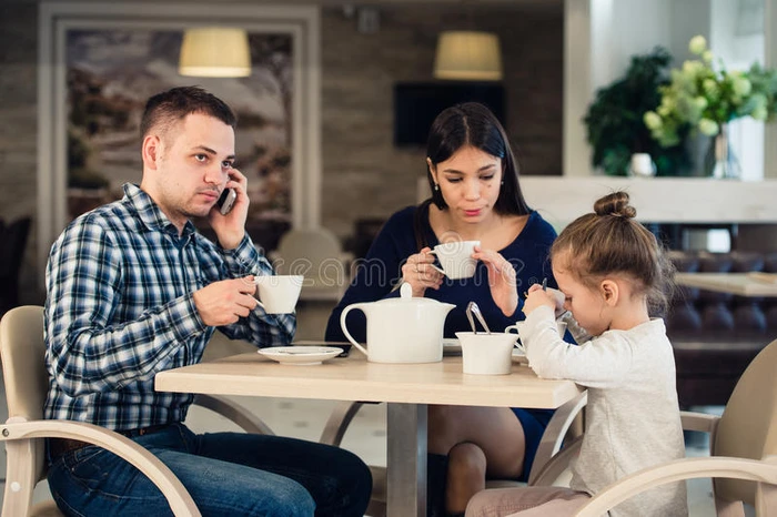 白种人的母亲和小女孩在餐馆里而他们忙碌的父亲在说手机